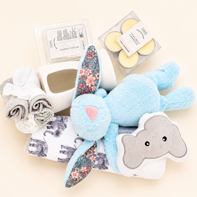Baby gift hamper featuring a blue bunny comforter, elephant-themed towel, scented wax melts, a ceramic burner and tealight candles.